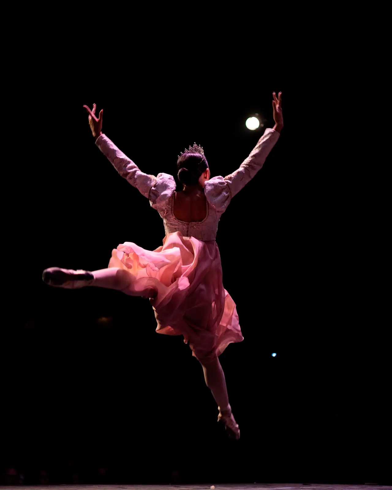 Bailarina con tiara y vestido rosa en pleno salto durante una función de ballet.